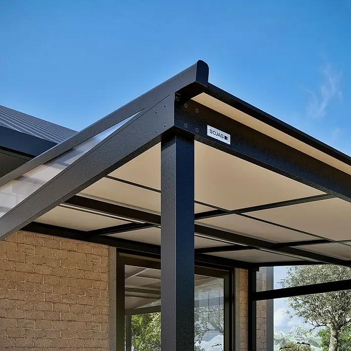 Sojag Terrassenüberdachung Budavar - Schwarz, Metall, L x B x H: 298,5 x 366,5 x 243 cm - Dunkle, texturierte Terrassenüberdachung mit hellem Dach und transparenten Seitenwänden, an ein Ziegelgebäude angebaut, unter blauem Himmel.