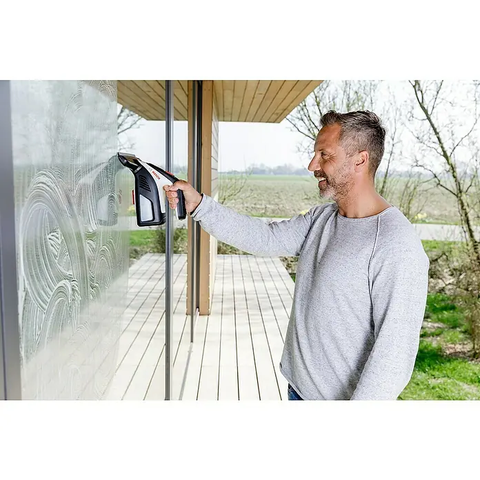 Akku-Fenstersauger - 3,6 V, 2 Ah, Behältervolumen: 100 ml - Mann lächelt beim Reinigen eines großen Fensters mit einem schwarz-weißen Bosch Fenstersauger.