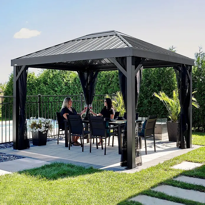 Sojag Standard-Pavillon Kiruna 1012 - Anthrazit, Metall, L x B x H: 299,3 x 364,3 x 297,6 cm - Dunkelgraues Metallpavillon mit schwarzen Netzvorhängen auf einer Terrasse, das zwei Frauen einen Essbereich im Freien bietet.