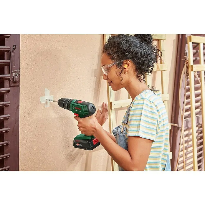 Bosch Power for All 18V Maschinen-Set 2-Tool-Kit EasyImpact + EasySaw - 18 V, 2 Akkus, 2 Ah, 5 -tlg. - Frau bohrt mit einem grün-schwarzen Bosch Akkubohrer ein Loch in eine helle Wand, mit Schutzbrille und Gehörschutz.