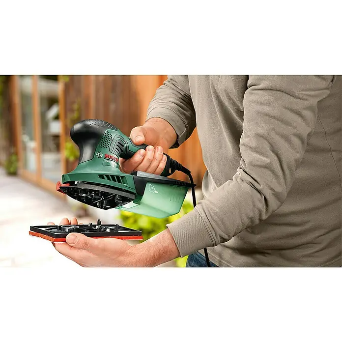 Bosch Elektro-Multischleifer PSM 200 AES SYSTEMBOX - 200 W, Leerlaufschwingzahl: 6.000 U/min - 26.000 U/min, Durchmesser Schwingkreis: 1,8 mm - Mann hält grünen Bosch Multischleifer (PSM 200 AES), wechselt die Schleifplatte. Das Gerät hat einen transparenten Staubbehälter.