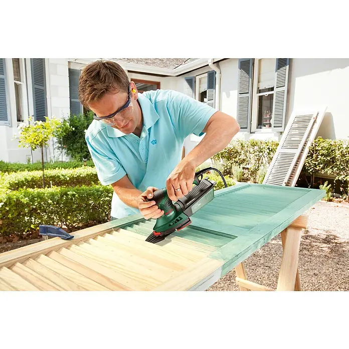 Bosch Elektro-Multischleifer PSM 200 AES SYSTEMBOX - 200 W, Leerlaufschwingzahl: 6.000 U/min - 26.000 U/min, Durchmesser Schwingkreis: 1,8 mm - Mann in blauem Hemd schleift grüne Holzjalousie mit grün-schwarzem Bosch Schleifer.
