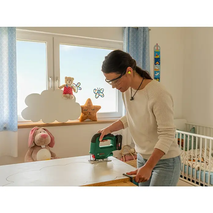 Bosch Power for All 18V Maschinen-Set 2-Tool-Kit EasyImpact + EasySaw - 18 V, 2 Akkus, 2 Ah, 5 -tlg. - Eine Frau schneidet mit einer grünen Bosch Akku-Stichsäge eine Wolkenform aus einem Holzbrett, mit Schutzbrille und Gehörschutz.