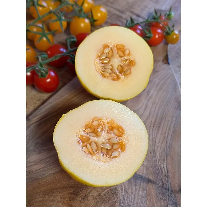 Wassermelone - Cucumis melo 'Marlene', Topfgröße: 12 cm - Zwei Hälften einer gelben Melone mit Kernen auf einem Holzbrett, mit Kirschtomaten im Hintergrund.