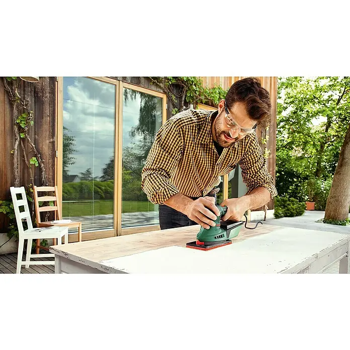 Bosch Elektro-Multischleifer PSM 200 AES SYSTEMBOX - 200 W, Leerlaufschwingzahl: 6.000 U/min - 26.000 U/min, Durchmesser Schwingkreis: 1,8 mm - Ein Mann schleift eine weiße Holzplatte im Freien mit einem grünen Bosch Schleifgerät.