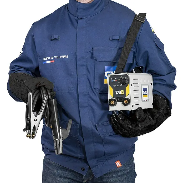Gys Schweißgerät GYSMI120A - Schweißstrom: 10 A - 120 A, L x B x H: 23 x 15 x 10 cm - Person in blauer Arbeitsjacke und Schweißhandschuhen trägt ein kompaktes GYSMI 120A Inverter-Schweißgerät mit Kabeln und Klemmen.