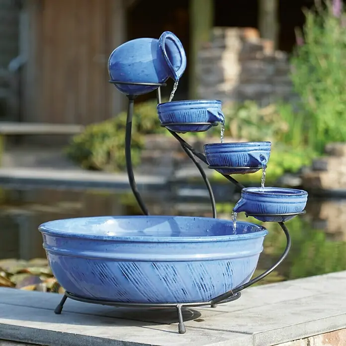 Ubbink Statuen-Zimmerbrunnenpumpe Eli indoor 600i - 9 W, Max. Förderhöhe: 130 cm - Blauer Kaskadenbrunnen mit vier Ebenen und Wasserfluss in einen großen Auffangbehälter auf einer Terrasse mit Teich im Hintergrund.