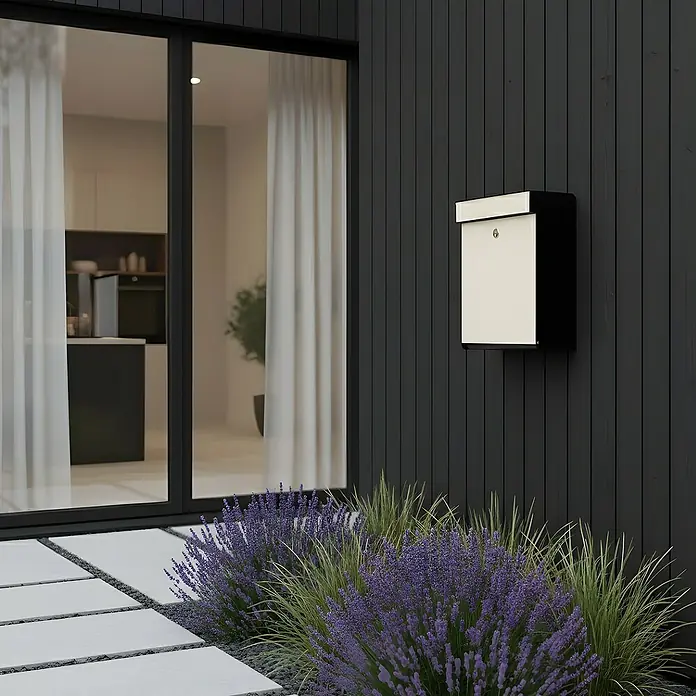 Allux Briefkasten Grundform - L x B x H: 180 x 345 x 440 mm, Schwarz / Cremeweiß, Stahl - Moderner schwarz-cremefarbener Briefkasten an einer dunklen vertikalen Wand eines Hauses, umgeben von Lavendel und Gräsern.