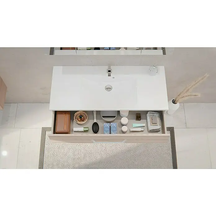Fackelmann Oxford Badmöbel-Set - Gesamtbreite Badmöbel: 120 cm, Braun Hell, Matt, 3-Teiliges Set mit Spiegelschrank - Blick von oben auf einen Waschtisch aus hellem Holz mit geöffneter Schublade, gefüllt mit Toilettenartikeln, Kosmetik und einer Haarbürste, unter einem weißen Waschbecken.