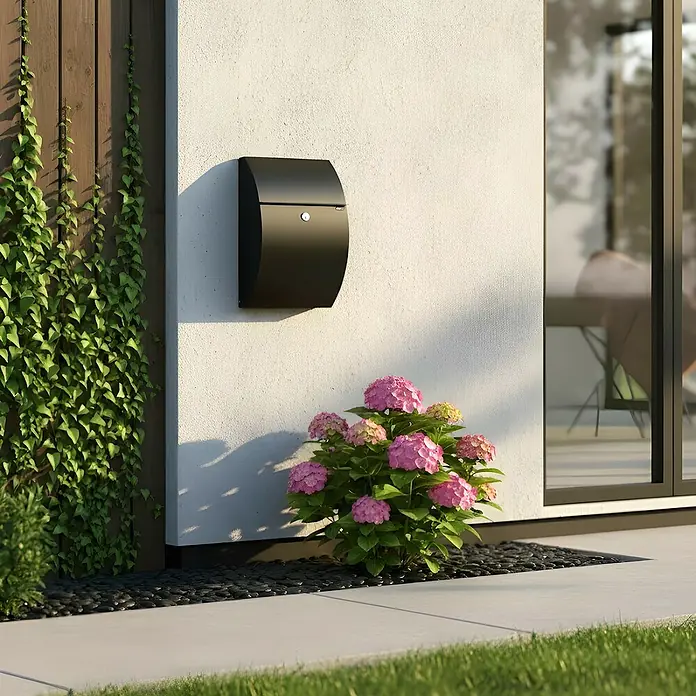Allux Briefkasten 7000 - L x B x H: 195 x 320 x 510 mm, Schwarz, Stahl - Eleganter schwarzer Briefkasten an einer weißen Hauswand, umgeben von grünen Kletterpflanzen und rosa Blumen.