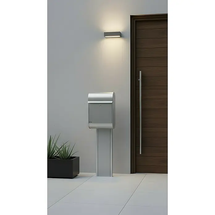 Allux Standbriefkasten 8900 - L x B x H: 160 x 340 x 500 mm, Silber, Stahl - Moderner silberner Briefkasten auf Ständer, an einem Eingang neben einer Holztür und einer Außenleuchte platziert.