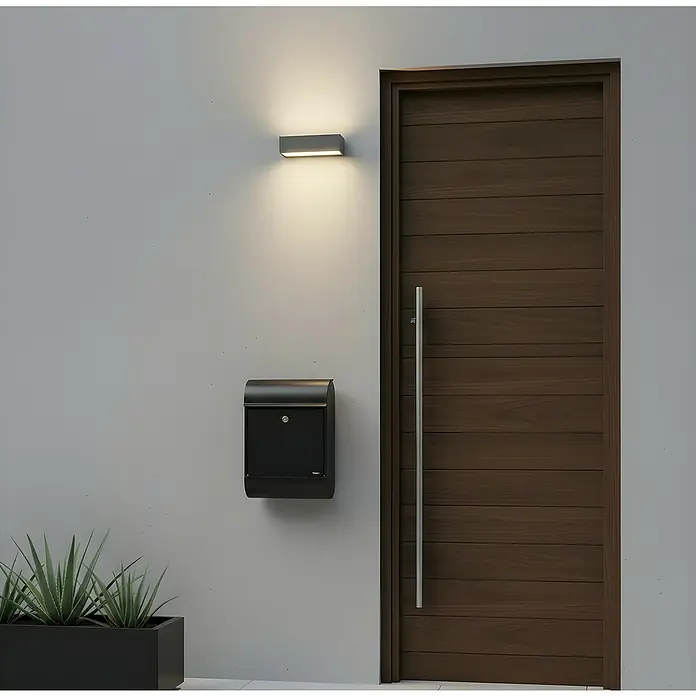 Allux Standbriefkasten 8900 - L x B x H: 160 x 340 x 500 mm, Schwarz, Stahl - Moderner Eingangsbereich mit grauer Außenleuchte, schwarzem Briefkasten, dunkler Holztür und einem schwarzen Pflanzgefäß mit Grünpflanzen.