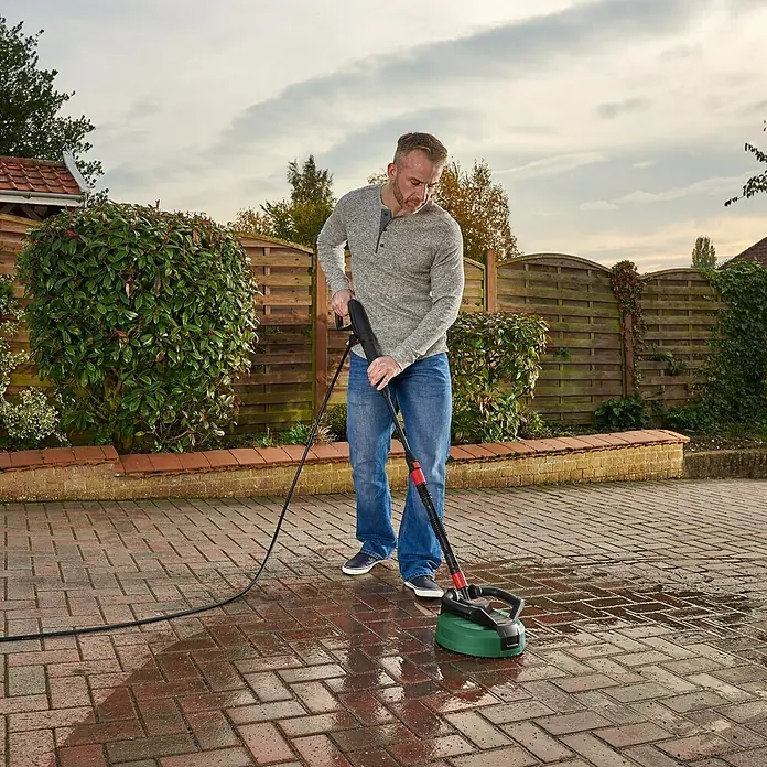 Bosch Flächenreiniger Multi Aquasurf 280 - Durchmesser: 280 mm, Passend für: Bosch Hochdruckreiniger Aquatak Serie - Ein Mann benutzt einen Terrassenreiniger, um eine Auffahrt zu reinigen. Das Werkzeug mit der grünen Bürste entfernt Schmutz von den Steinen.