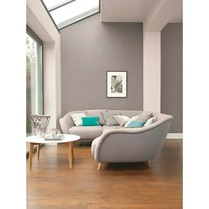 Bruguer Colores del Mundo Pintura para paredes - Nepal matiz de beige, Mate, 4 l - Un salón luminoso con un sofá esquinero gris con cojines turquesa y una mesa de centro blanca sobre un suelo de madera.