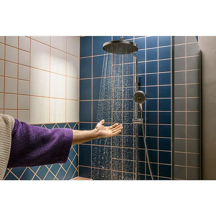 Hansgrohe Duschsystem Activera S - Lochabstand: 138 mm - 162 cm, Anzahl Strahlarten: 2 Stk., Chrom - Hand im lila Bademantel testet Wasser aus schwarzem Duschkopf in einer modernen blau-weiß gefliesten Dusche.