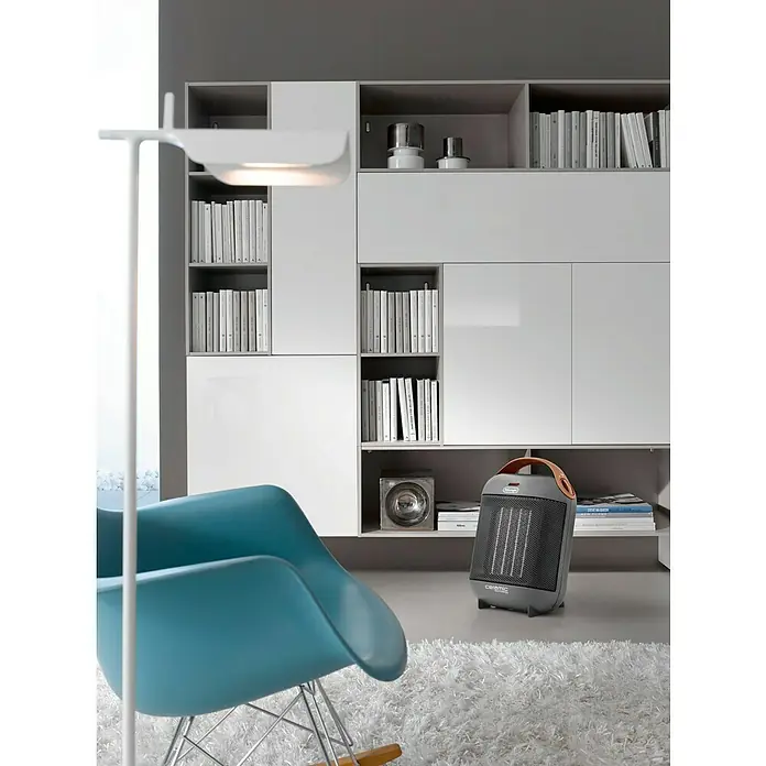 De'Longhi Calefactor de aire Capsule - 900 W - 1.800 W, Gris oscuro - Un calefactor cerámico gris con asa está sobre una alfombra blanca frente a una estantería moderna, brindando calor y confort al espacio.