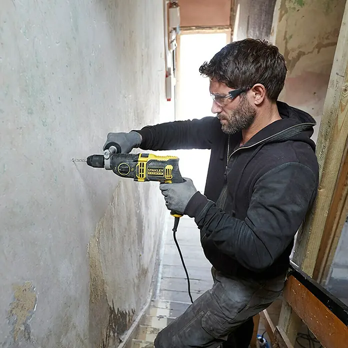 Stanley FatMax Taladro percutor FMEH850K-QS - 850 W, Portabrocas de sujeción rápida - Un hombre está usando un taladro Stanley Fatmax para hacer un agujero en una pared. Se están usando equipos de seguridad.