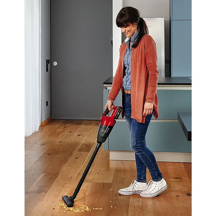 Einhell Power X-Change 18V Akku-Handstaubsauger TE-VC 18 Li-Solo - 18 V, Ohne Akku - Eine Frau saugt Krümel mit einem Einhell Akku-Staubsauger auf einem Holzboden auf. Das Gerät ist rot und schwarz.