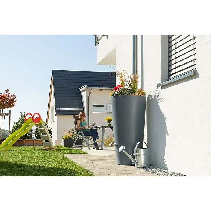 Arves Regenspeicher Rainbowl Flower - 150 l, Betongrau - Ein grauer Pflanzkübel aus Kunststoff mit Pflanzen, neben einer silbernen Gießkanne, an einer weißen Hauswand.