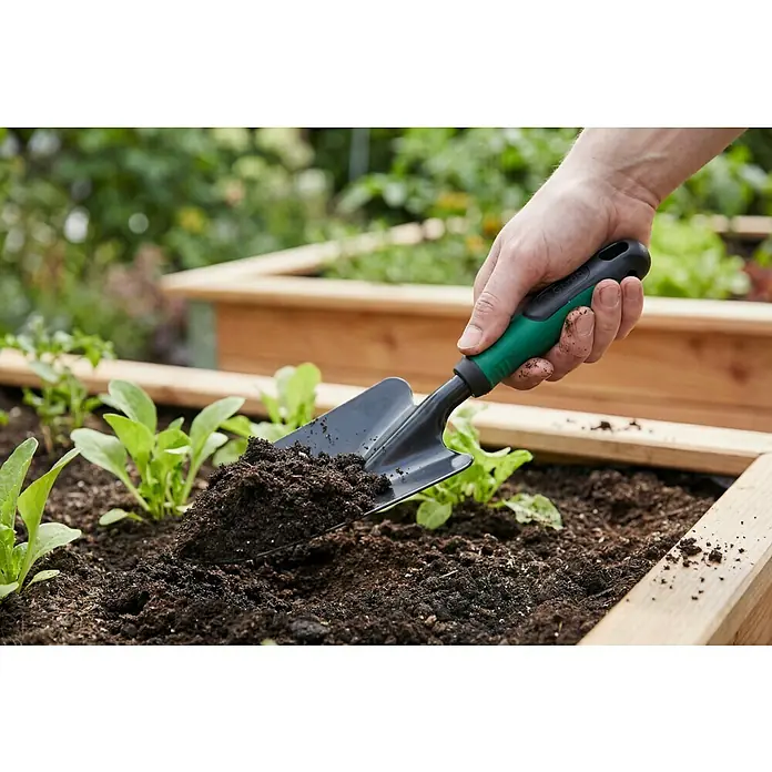 Gardol Hand-Blumenkelle - 10 cm, Stahl, Kurzstiel - Hand hält eine grün-schwarze Gartenschaufel, die dunkle Erde in einem Hochbeet mit jungen Pflanzen schaufelt.