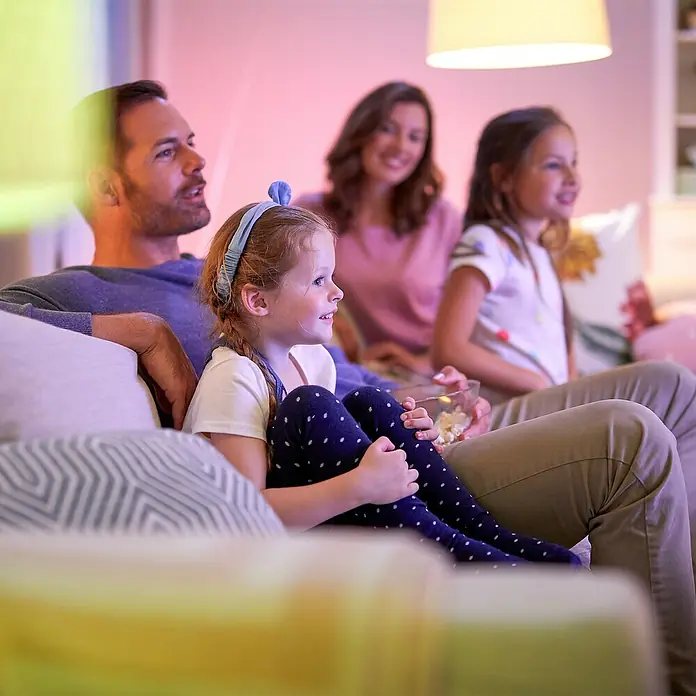 Philips Wiz Smart-LED-Lampe matter - E27, Dimmbar, RGBW, 1.521 lm, 13 W, 1 Stk. - Familie auf Couch schaut Unterhaltung, beleuchtet von farbigem Smart Lighting, schafft gemütliche Atmosphäre.