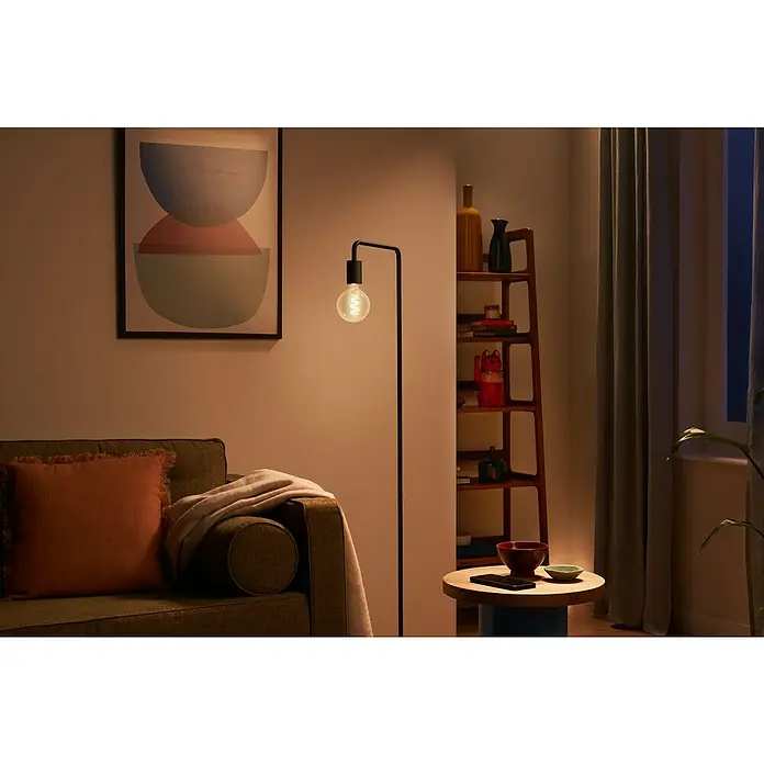 Philips Wiz Smart-LED-Lampe Globe - E27, Dimmbar, RGBW, 470 lm, 6,3 W, 1 Stk. - Eine schwarze Stehlampe mit spiralförmiger Glühbirne beleuchtet gemütlich ein Wohnzimmer neben einem Sofa.