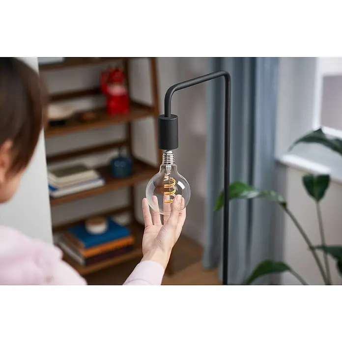 Philips Wiz Smart-LED-Lampe Globe - E27, Dimmbar, RGBW, 470 lm, 6,3 W, 1 Stk. - Hand installiert eine klare Glühbirne mit Spiralfilament in eine moderne schwarze Stehlampe.