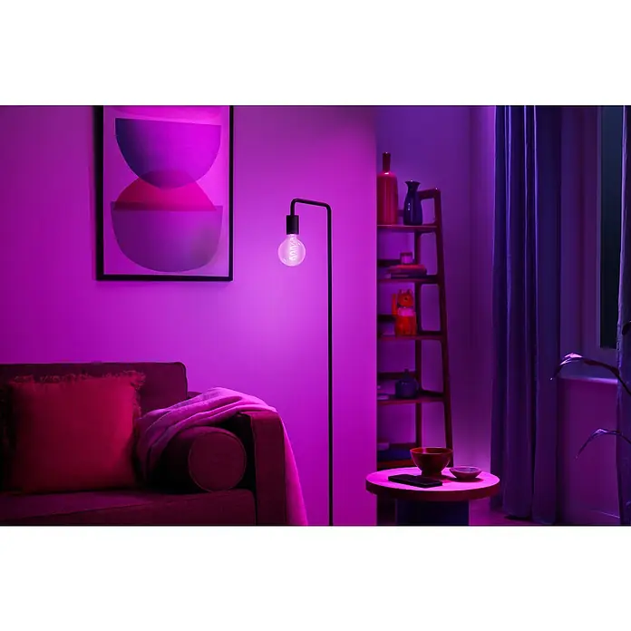 Philips Wiz Smart-LED-Lampe Globe - E27, Dimmbar, RGBW, 470 lm, 6,3 W, 1 Stk. - Eine smarte Glühbirne mit sichtbarem Filament in einer Stehlampe taucht ein modernes Wohnzimmer in lebhaftes Lila.