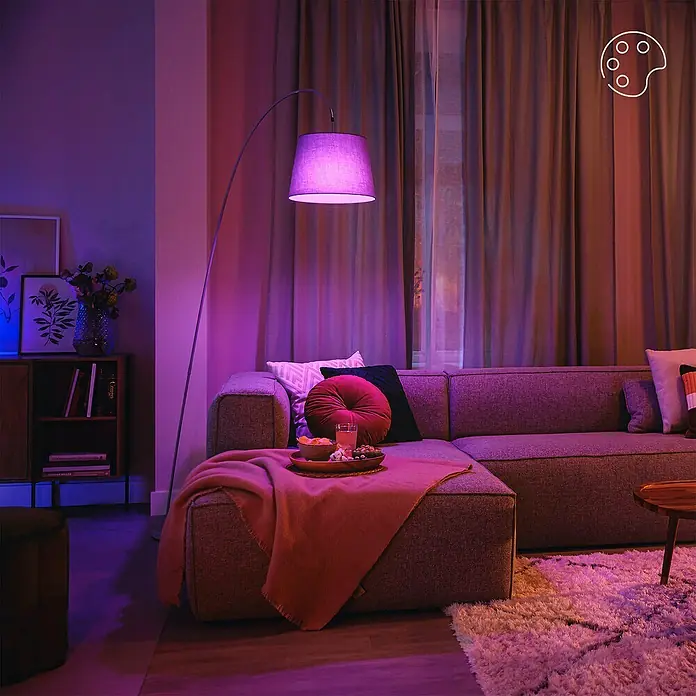 Philips Wiz Smart-LED-Lampe matter - E27, Dimmbar, RGBW, 806 lm, 8,5 W, 1 Stk. - Eine lila Stehlampe beleuchtet ein gemütliches Wohnzimmer und schafft eine entspannte Abendatmosphäre um ein Sofa mit Snacks.