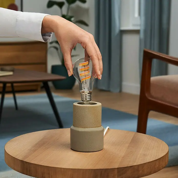 Philips Wiz Smart-LED-Lampe - E27, Dimmbar, RGBW, 470 lm, 6,3 W, 1 Stk. - Hand schraubt eine LED-Glühbirne mit Spiralfilament in einen Betontischlampensockel.