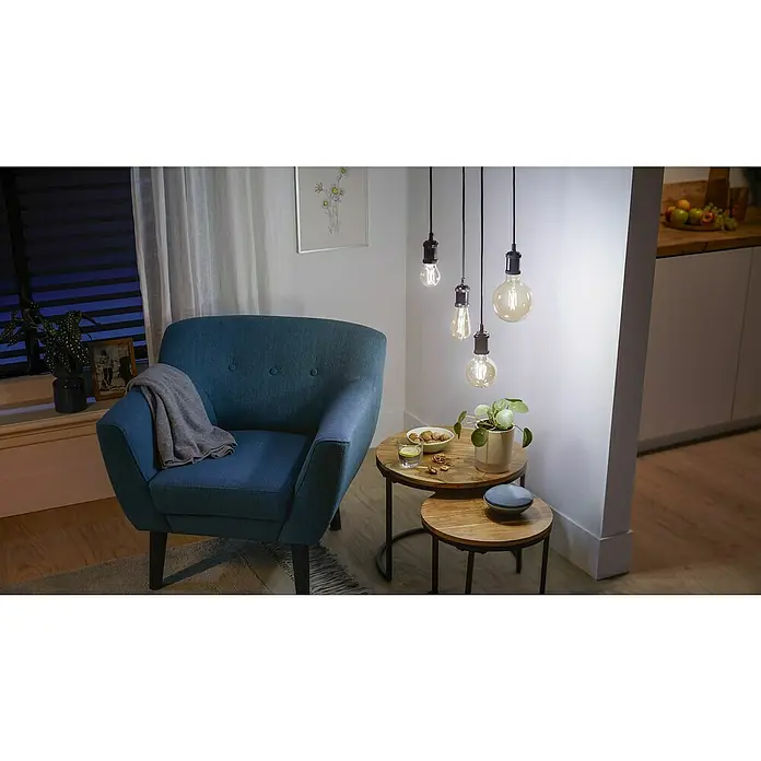 WiZ Smart-LED-Lampe ST64 - E27, Dimmbar, Warmweiß, 640 lm, 7 W, 1 Stk. - Vier hängende Glühbirnen im Vintage-Stil mit warmem Licht beleuchten ein gemütliches Wohnzimmer mit blauem Sessel und Holz-Beistelltischen.