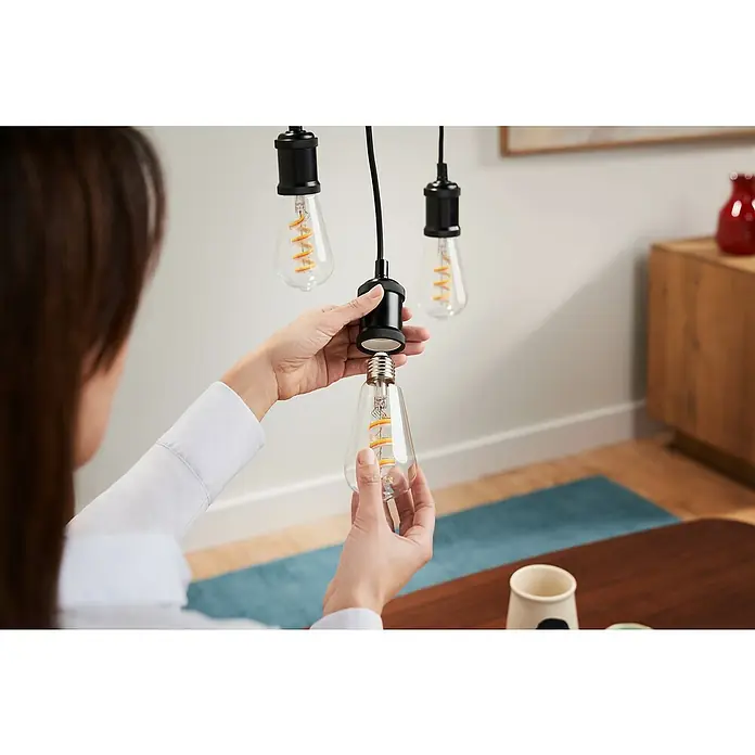 Philips Wiz Smart-LED-Lampe Edison - E27, Dimmbar, RGBW, 470 lm, 6,3 W, 1 Stk. - Eine Person schraubt eine klare LED-Glühbirne mit Spiralfilament in eine schwarze Pendelleuchtenfassung.