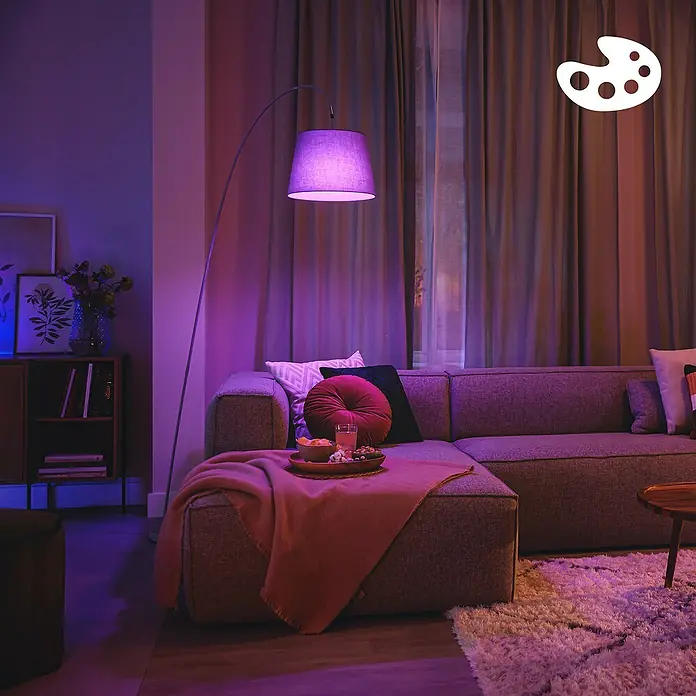 Philips Wiz Smart-LED-Lampe - E27, Dimmbar, RGBW, 2.452 lm, 18,5 W, 1 Stk. - Wohnzimmer mit Bogenstehlampe, die violettes Licht abstrahlt. Ein graues Ecksofa ist mit Kissen und einem Snacktablett bestückt.