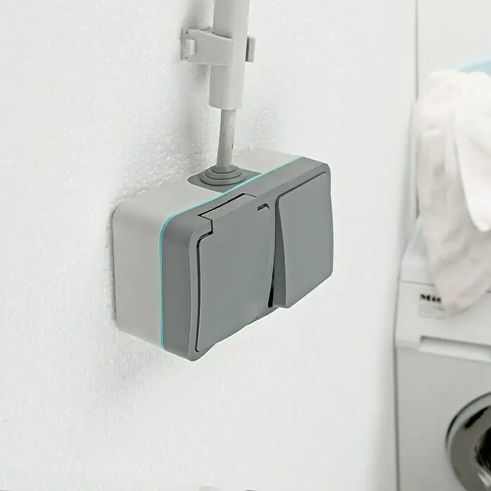 Voltomat Blueline Feuchtraum-Schalterkombination - Grau, Ausrichtung Schalterprogramm: Horizontal, Aufputzmontage, IP54 - Eine graue Aufputz-Steckdose mit Klappdeckel und seitlicher blauer Akzentlinie ist an einer weißen Wand montiert, Kabel führt nach oben.