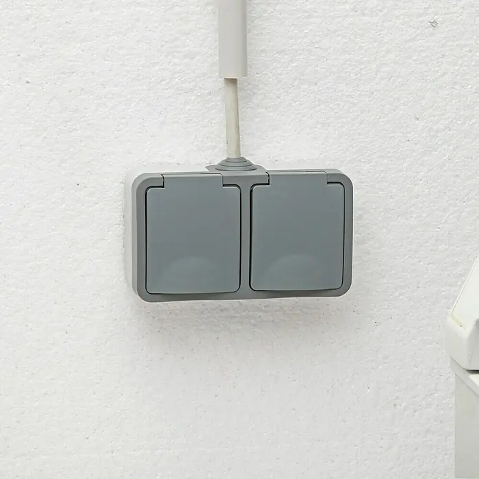 Voltomat Blueline Feuchtraum-Steckdose - 2-fach, Horizontal, IP54, Grau - Eine graue Doppelsteckdose mit Klappdeckel ist an einer weißen Wand montiert und durch ein Kabel mit Strom versorgt.