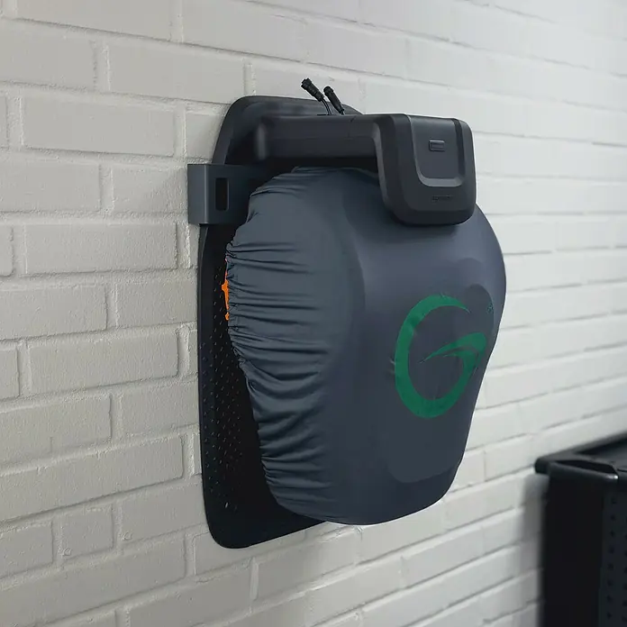 Grimsholm Green Abdeckhaube für Rasenmäher - L x B: 290 x 240 mm, Gängige Mähroboter-Modelle - An der Wand montierte schwarze Ladestation mit grauem, grün logo-bedrucktem Gerätebezug. Kabel ragen oben heraus.