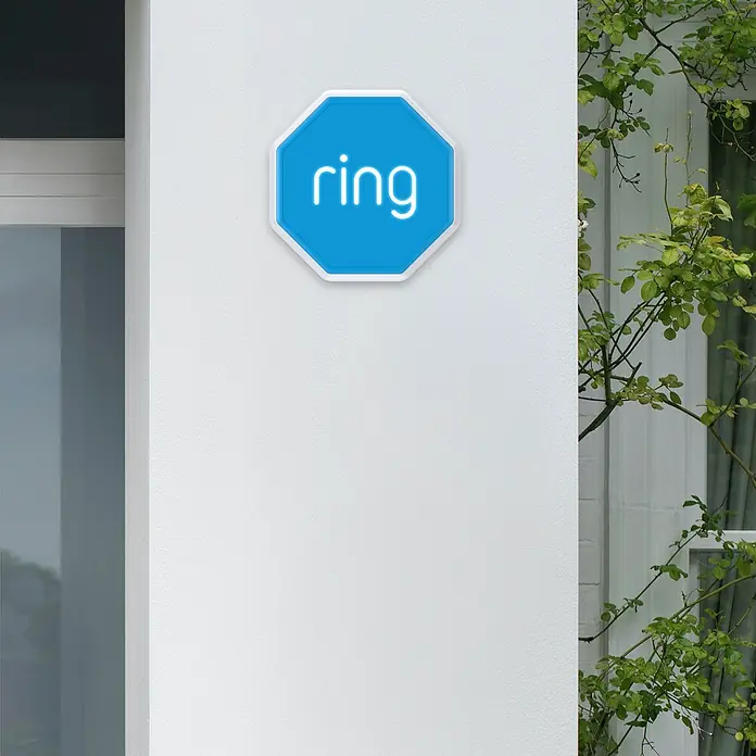 Ring Alarmsirene Outdoor - 100 dB, Batteriebetrieben - Blaues achteckiges Ring-Sicherheitsschild mit weißem Logo, an weißer Außenwand neben einem Fenster montiert.