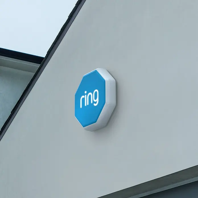 Ring Alarmsirene Outdoor - 100 dB, Batteriebetrieben - Weiße, achteckige Ring Außenalarmsirene mit blauem Logo-Panel, an einer Hauswand montiert.