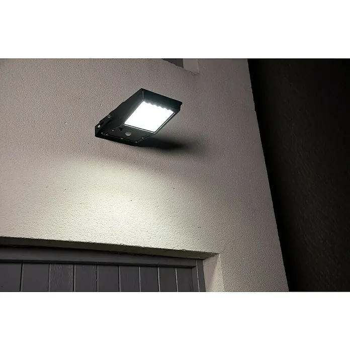 Osram LED-Sensor-Außenwandleuchte Endura Flood - 10 W, 19,4 x 6,7 x 31,3 cm, Kaltweiß - Schwarzer LED-Außenstrahler mit Bewegungssensor, an einer strukturierten Wand montiert, strahlt helles Licht aus.
