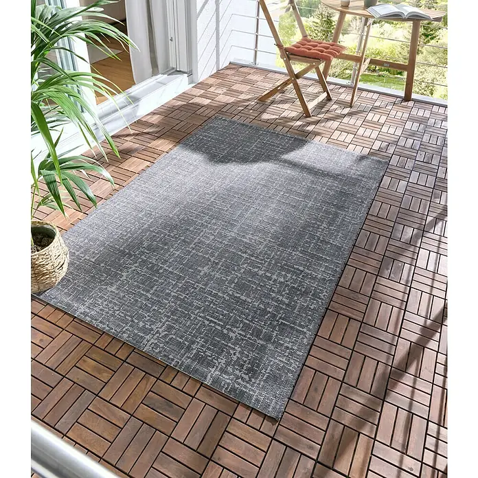 Alfombra exterior Newberry Antracita - Antracita, 230 x 160 cm, 100% Polipropileno - Alfombra exterior gris con un patrón sutil, colocada sobre un suelo de lamas de madera en un balcón.