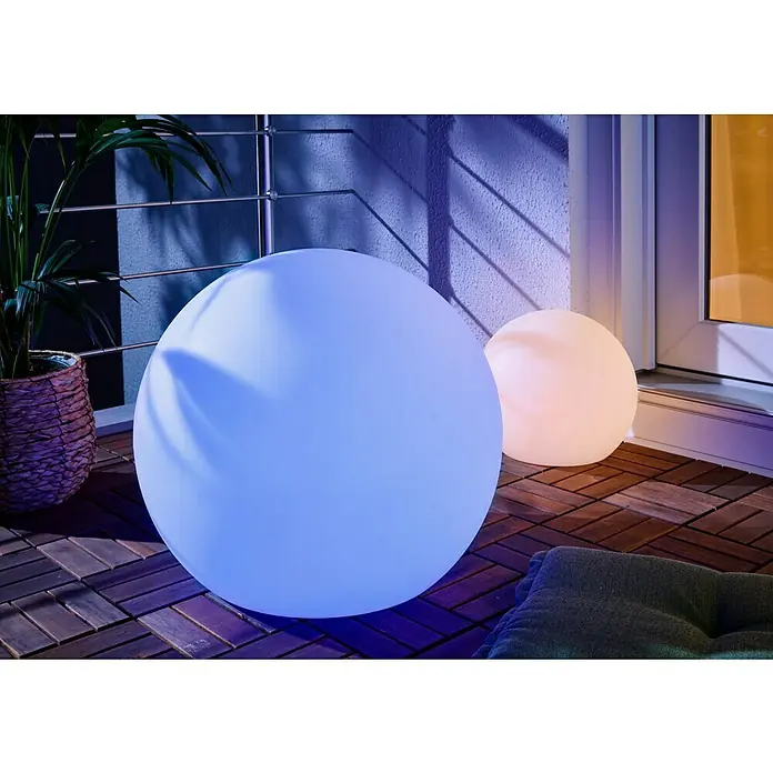 New Garden Solar-Dekoleuchte Buly - Durchmesser: 50 cm, IP65 - Zwei leuchtende kugelförmige Außenlampen, eine große blaue und eine kleinere orange, auf einem gefliesten Holzbalkon mit Pflanze.