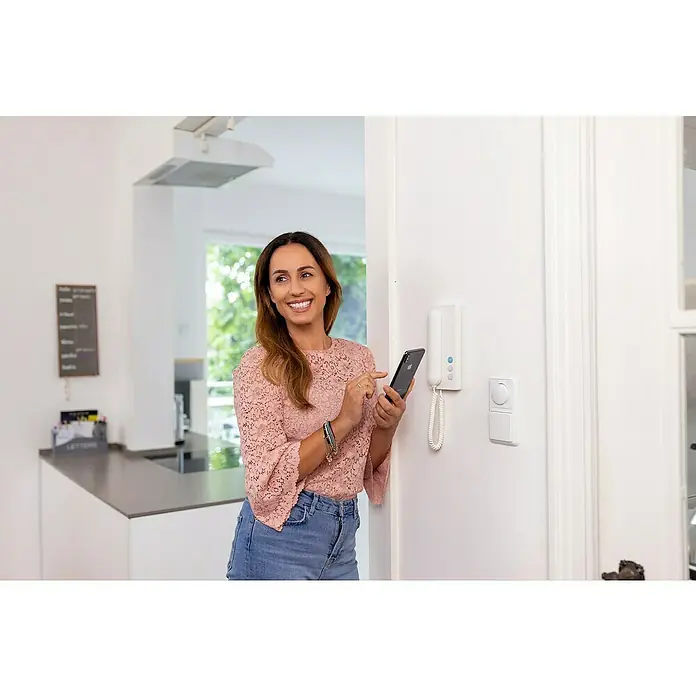 Siedle Türsprechanlage Compact SET CA 812-4 E/W - 4 Personen-Haushalt, Weiß, Silber - Frau bedient ein weißes, wandmontiertes Smart-Home-Bedienfeld und eine Gegensprechanlage mit ihrem Smartphone.