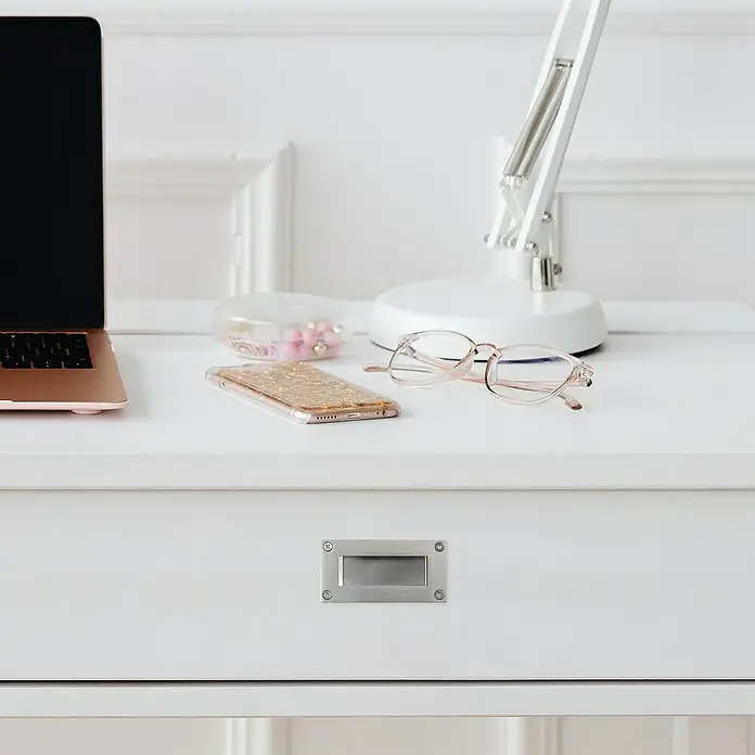Rei Möbelgriff - Lochabstand: 96 mm, Sonstige, Edelstahl - Weißer Schreibtisch mit roségoldenem Laptop, goldglitzerndem Telefon, rosa Brille und weißer Lampe. Ein rechteckiger Metallschubladengriff am Schubkasten.