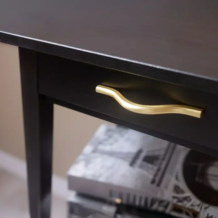 Rei Möbelgriff - Lochabstand: 96 mm, Aluminium, Gold - Nahaufnahme einer schwarzen Holzschublade mit goldenem, geschwungenem Griff. Darunter sind verschwommene Bücher zu sehen.