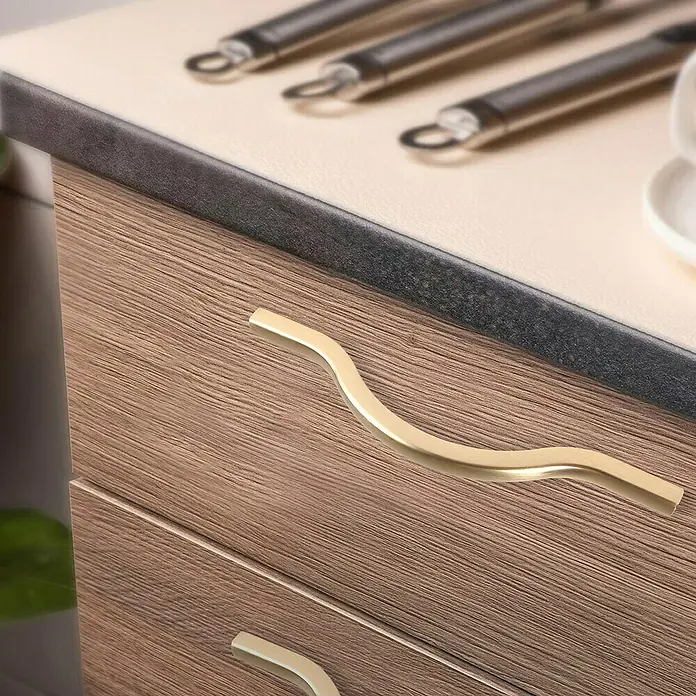 Rei Möbelgriff - Lochabstand: 96 mm, Aluminium, Gold - Goldfarbener, wellenförmiger Schrankgriff an einer Holzschublade in Küchenumgebung.