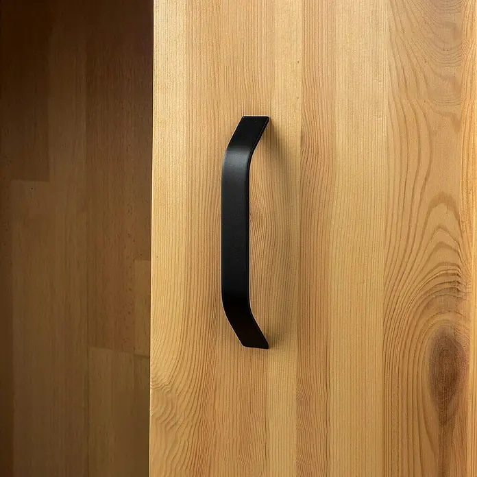 Rei Möbelgriff - Lochabstand: 160 mm, Aluminium, Schwarz, Gebogen - Schwarzer matter Griff an einer hellen Holztür, mit sichtbarem dunklem Schrankinneren.