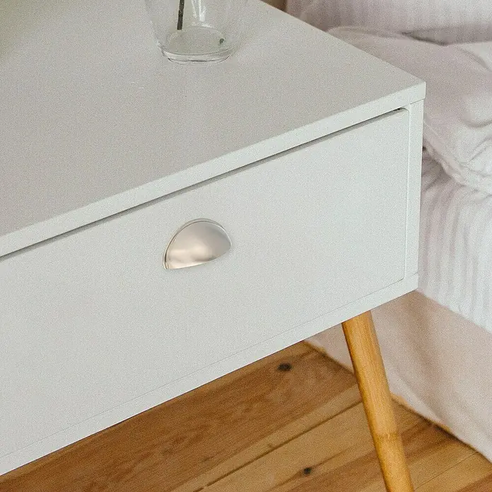 Rei Möbelgriff - Lochabstand: 64 mm, Sonstige, Nickel, L x B x H: 80 x 29 x 25 mm - Weißer Nachttisch mit silbernem Muschelgriff, Holzbeinen, neben einem Bett, mit einem Glas darauf.