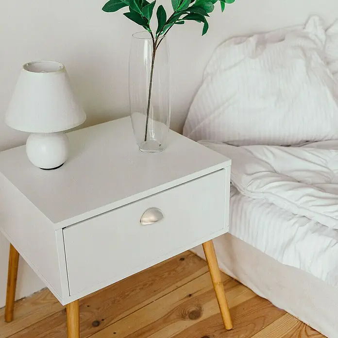 Rei Möbelgriff - Lochabstand: 64 mm, Sonstige, Nickel, L x B x H: 80 x 29 x 25 mm - Ein weißer Nachttisch mit Holzbeinen und silbernem Griff, auf dem eine Lampe und Vase stehen, neben einem weißen Bett.