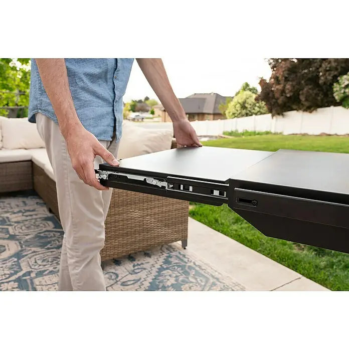 Blackstone Plancha Grill - Superficie de la parrilla (An x Pr): 91 x 55 cm, Negro, Número de quemadores: 4 ud. - Las manos de una persona sostienen una extensión de mesa negra, revelando su mecanismo deslizante metálico en un patio.