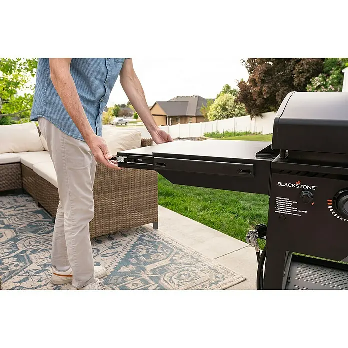 Blackstone Plancha Grill - Superficie de la parrilla (An x Pr): 91 x 55 cm, Negro, Número de quemadores: 4 ud. - Hombre ajustando el estante lateral de una plancha Blackstone negra en un patio.